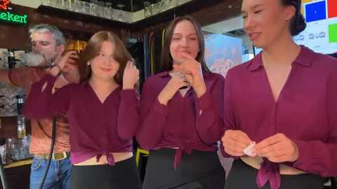 Media: Video of four women in maroon tops and black pants, smiling, interacting, in a lively bar setting. Background includes a bar counter, glasses, and colorful decor.