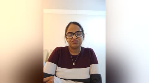 Media: A video of a woman with medium-dark skin, wearing glasses, a maroon and white striped sweater, and a necklace, sitting indoors with a soft, warm light illuminating her from the left.