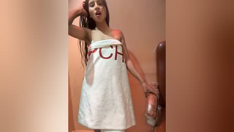 Media: Video of a young woman with long brown hair, wearing a white towel with red \"FUCK\" text, standing in a dimly lit bathroom, holding a clear glass dildo.