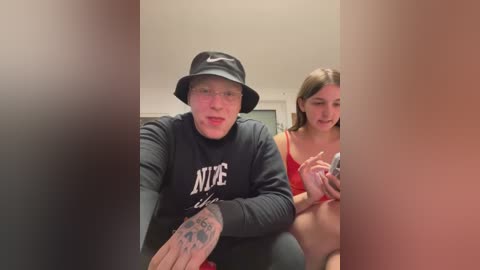 Media: Video of a young man in a black Nike sweatshirt and bucket hat, with a tattooed hand, sitting next to a young woman in a red bra, both smiling.