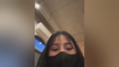 Media: A blurred video of an Asian woman with dark hair, wearing a black mask, standing in a beige room with a blue window and an air conditioner.