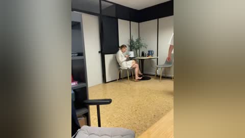 Media: A video of a modern office with a woman in a white shirt and shorts, sitting at a desk with a laptop, surrounded by beige walls and wooden floors.