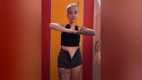 Media: Video of a fit, tattooed, blonde woman in black sports bra and shorts, standing in a red and yellow-striped room, striking a pose against a wall.