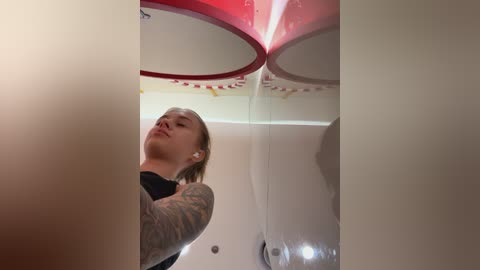 Media: Video of a young woman with blonde hair and a tattooed arm, wearing headphones, inside a white, futuristic, circular room with red accents and circular lights.