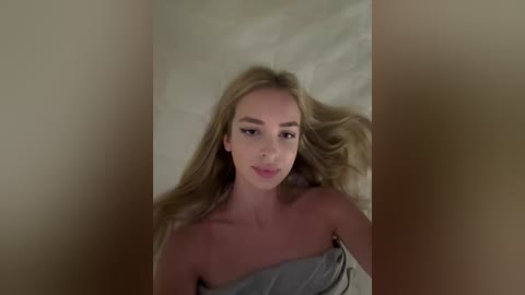 Media: A video of a young woman with long blonde hair lying on a beige bed, wearing a gray off-shoulder top, with a blurred background.