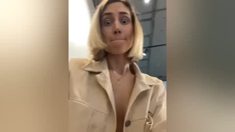 Media: Video of a woman with short blonde hair, wearing a beige jacket that's unbuttoned, revealing a cleavage, in a dimly lit room with white walls and a ceiling.