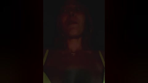 Media: Video of a woman with medium brown skin, wearing a green top, standing in a dark room, with a blurred background.