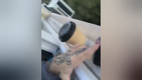 Media: A blurred video shows a tattooed person, possibly male, holding a coffee cup, in a grassy outdoor setting with a white vehicle and green foliage in the background.