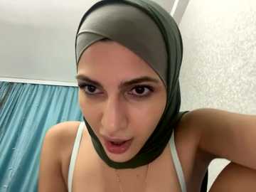 Media: Video of a young woman with olive skin, wearing a green hijab and white tank top, looking seductively into the camera with parted lips. Background features a light-colored wall and teal curtains.