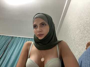 Media: Video of a light-skinned woman with a green hijab and beige bra, wearing a pendant necklace, in a room with teal curtains and patterned wallpaper.