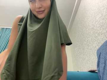 Media: A video of a young woman with fair skin and short dark hair, wearing a green hijab and sleeveless top, standing in a bathroom with teal curtains and textured wallpaper.