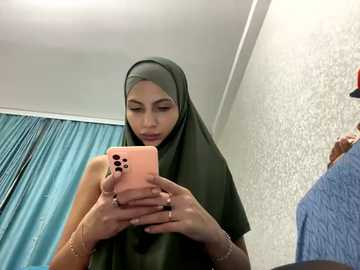 Media: Video of a young woman with medium skin tone, wearing a dark green hijab and sleeveless top, taking a selfie in a modern room with white walls and blue curtains.