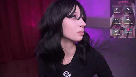 Media: Video of a pale-skinned, slender woman with long black hair, wearing a black top, looking to the right. Background shows a purple-lit room with a digital display screen showing a live stream.