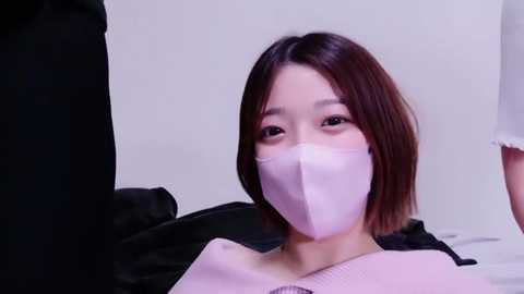 Media: Video of an East Asian woman with shoulder-length brown hair, wearing a light pink surgical mask and white medical gown, lying on a white bed in a sterile room.