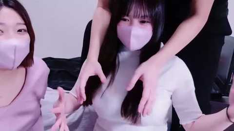 Media: Video of three Asian women with long, straight black hair, wearing face masks and white turtlenecks, seated on a black couch. One woman is being held by two others, hands on her shoulders. Background is a plain, light-colored wall.