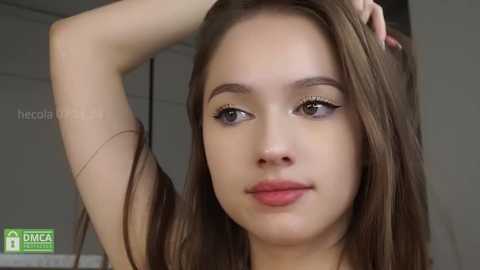 Media: Video of a young woman with fair skin, straight brown hair, and green eyes. She has a thin build, small breasts, and is wearing a black top. The background is a blurred indoor setting.