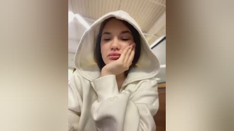 Media: Video of a young woman with fair skin, long dark hair, wearing a white hoodie, resting her head on her hand, with a blurred background featuring a ceiling and wall.