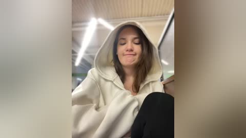 Media: A video captures a young woman with fair skin and straight, dark brown hair, wearing a white hoodie and black pants, smiling slightly in a brightly lit room with fluorescent lights and a beige ceiling.