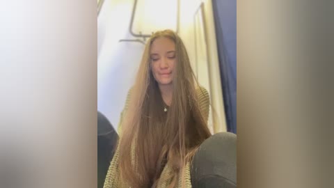 Media: Video of a young woman with long, straight brown hair, wearing a beige knitted sweater and dark jeans, sitting on airplane seats with a relaxed, contemplative expression.