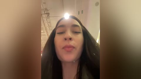 Media: Video of a young woman with long, dark hair, closed eyes, and puckered lips, taken from a low angle in a dimly lit hallway with a ceiling fan.