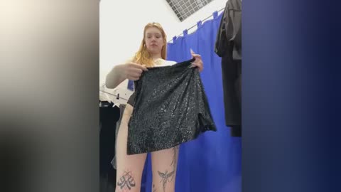 Media: A video of a fair-skinned, blonde woman with tattoos on her thighs, holding a black glittery skirt in a dressing room with blue curtains and a gray grid ceiling.