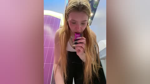 Media: Video of a young woman with long blonde hair, wearing a black top and pants, licking a pink lollipop, standing in a modern, colorful room with purple and blue tiles.