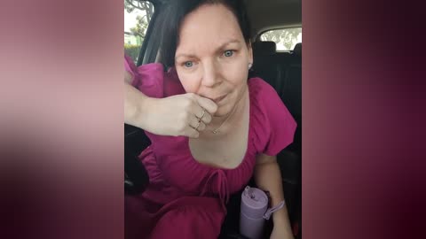 Media: A video of a middle-aged woman with fair skin and short dark hair, wearing a pink dress, resting her chin on her hand in a car seat. The background shows the car interior.