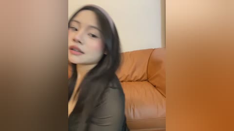 Media: A blurry video of an East Asian woman with long black hair, wearing a sheer black top, sitting on a brown leather couch against a beige wall.