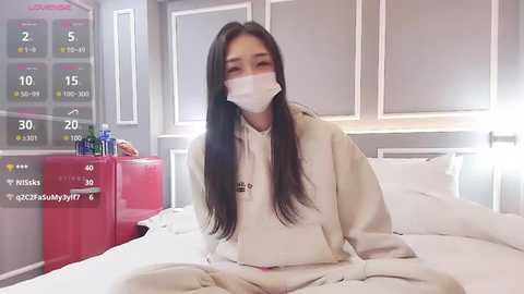 Media: Video of an Asian woman with long black hair, wearing a white surgical mask and beige hospital gown, sitting on a hospital bed with white linens, in a modern, bright room with a red cabinet and a digital thermometer.