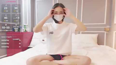 Media: Video of an Asian woman with short black hair, wearing a white mask, sitting cross-legged on a bed in a modern bedroom, adjusting her mask.