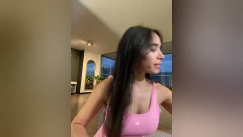 Media: Video of a young woman with long dark hair, wearing a pink tank top, doing yoga in a modern, minimalist living room with large windows.