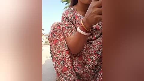 Media: Video of a dark-skinned woman in a floral-patterned sari, wearing a white bracelet, crouching in a bright, sunny outdoor setting.