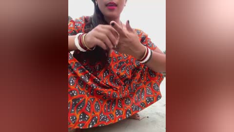 Media: Video of a woman with medium-brown skin, wearing a red and black patterned dress, bending down, hands near her face, against a plain white background.