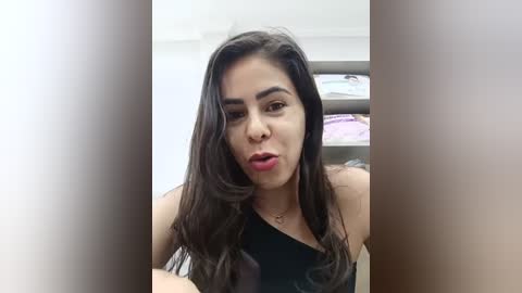 Media: A video of a young woman with long, dark hair, wearing a black off-shoulder top, making a playful face. She has medium skin tone and is in a brightly lit room with shelves in the background.