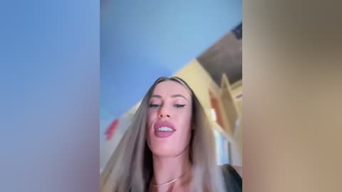 Media: Video of a young, light-skinned woman with long, straight, platinum blonde hair, wearing makeup with pink lipstick, captured from a low angle, showing her upper torso and face. Background shows a blurred, light-colored interior with a window.