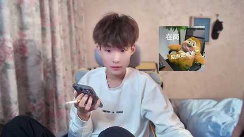 Media: Video of a young Asian boy in a white sweatshirt, holding a smartphone. Background features a floral curtain, beige wall, and a stuffed toy duck.