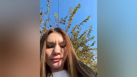 Media: Video of a young woman with light skin and long, straight, brown hair, wearing a white top, smiling, against a bright blue sky with green trees in the background.