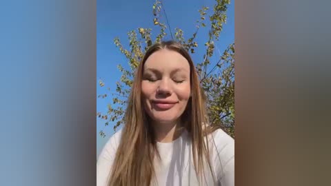 Media: Video of a young Caucasian woman with long, straight blonde hair, smiling with closed eyes, wearing a white top, standing outdoors with a clear blue sky and green leafy branches in the background.