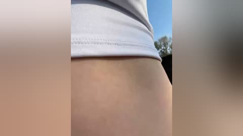 Media: A close-up video of a person's bare buttocks and upper thigh, showing skin texture, subtle folds, and a white garment partially covering the waist. The background features a blurred, outdoor setting with a clear blue sky and trees.