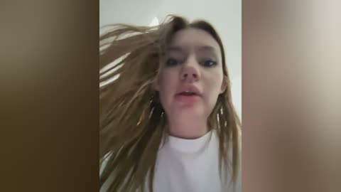 Media: Video of a young woman with fair skin and long, light brown hair, wearing a white turtleneck, captured from a low angle, with her hair blowing, creating a dynamic and expressive effect.