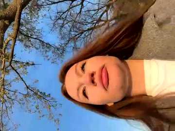 Media: A video of a young woman with long brown hair, fair skin, and light makeup, smiling outdoors. The background features a clear blue sky and sparse tree branches.