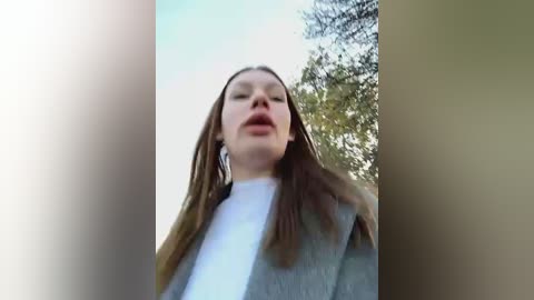 Media: Video of a woman with long brown hair and fair skin, wearing a white turtleneck and gray blazer, standing in a wooded area with green foliage and blue sky in the background.