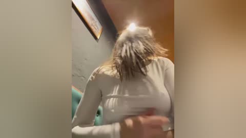 Media: Video of a woman with wet, tousled hair, wearing a white, long-sleeve top, captured in a bathroom with teal walls and a framed picture.