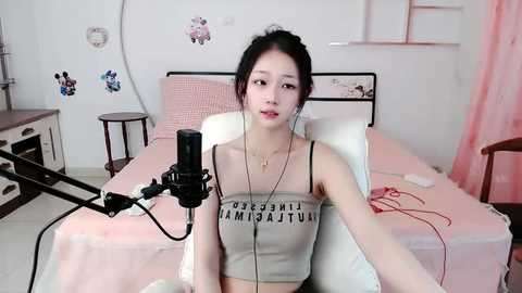 Media: Video of an Asian woman with pale skin, black hair, wearing a crop top with \"Hollywood\" in bold letters, recording herself on a microphone in a pastel pink bedroom.