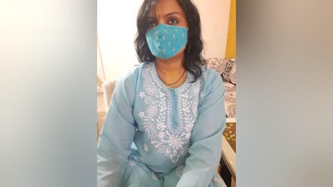 Media: Video of a South Asian woman with medium-brown skin and black hair, wearing a light blue traditional outfit with intricate embroidery, a blue face mask, and sitting in a modern living room with a couch and patterned cushions.