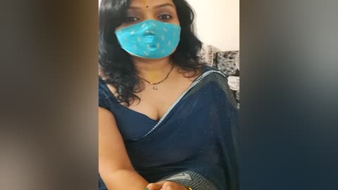 Media: Video of a South Asian woman with medium skin tone, wearing a blue saree and a turquoise mask, sitting indoors, her face partially obscured by a blurry foreground.