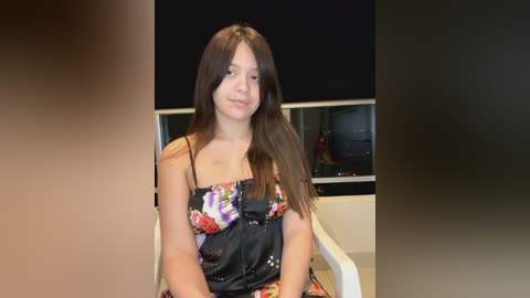 Media: Video of a young Caucasian girl with long brown hair, wearing a colorful floral dress, sitting in a modern, minimalist room with a black wall and white furniture.