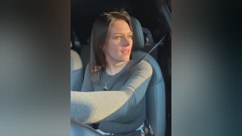 Media: Video of a Caucasian woman with light skin and shoulder-length brown hair, wearing a grey top and seatbelt, looking concerned while driving in a car.