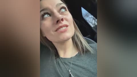 Media: Video of a young Caucasian woman with shoulder-length blonde hair, wearing a grey top, looking upward with a slight smile. The background features a blurred, dark car interior with a hint of tree branches outside.