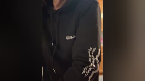 Media: Video of a person wearing a black hoodie with white text on the left sleeve and a small logo on the chest. The background is dimly lit, creating a moody atmosphere.